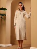 A woman modeling a long-sleeved, button-front midi shirt dress in a light beige linen-like fabric, standing in a warm beige architectural setting with arches and a plant.
