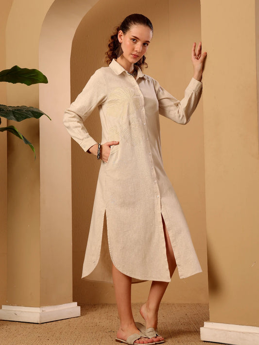 A woman modeling a light beige long-sleeve button-up shirt dress with a pointed collar, front pockets, and a split hem, posed in a warm beige architectural setting.