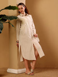 A woman models a long-sleeved, light beige linen shirt dress with a high slit, standing in a warm beige room next to a potted plant.