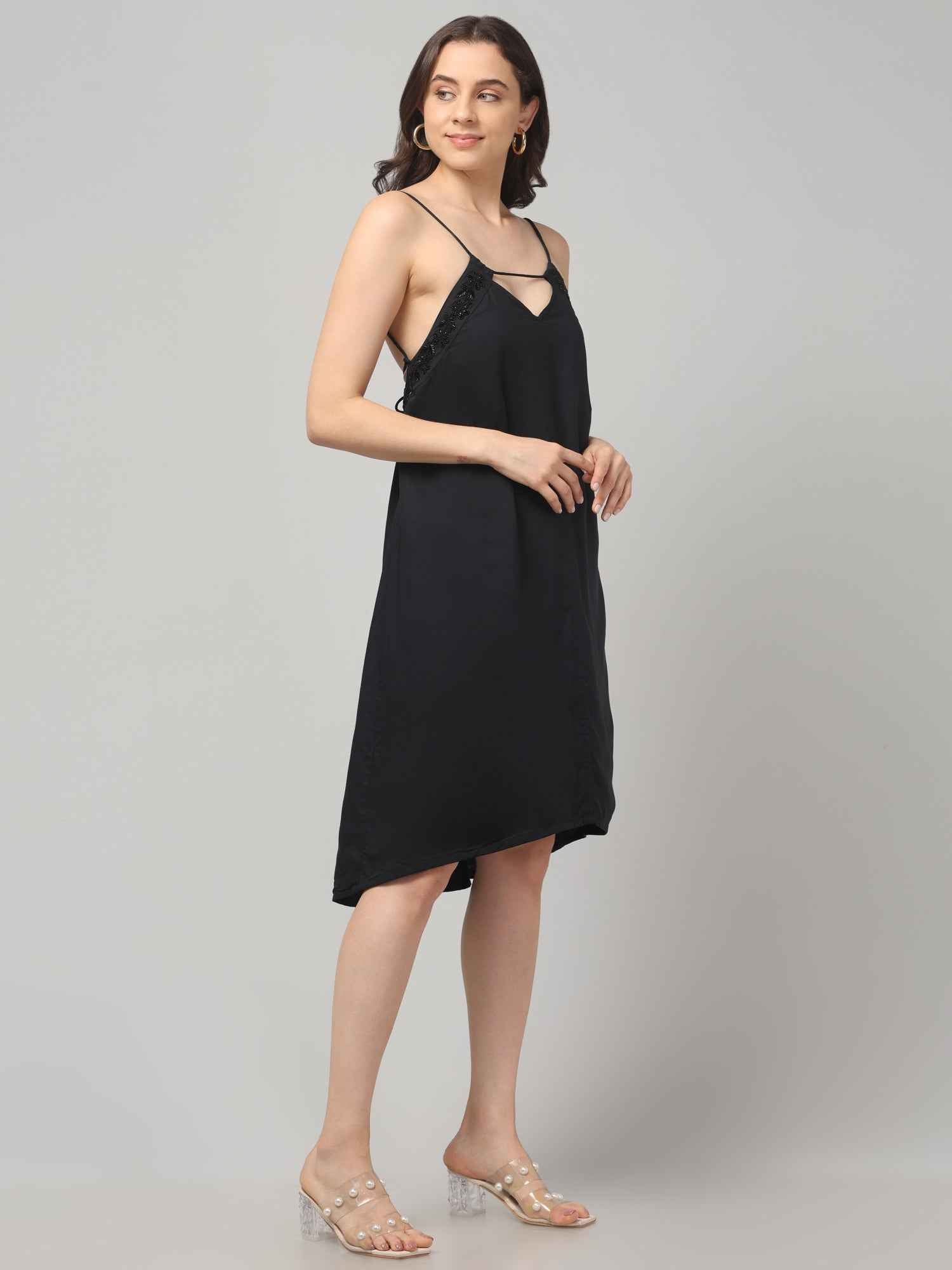 Side profile of a women wearing a black resort wear cocktail dress with noodle strap and black embellishment on neckline