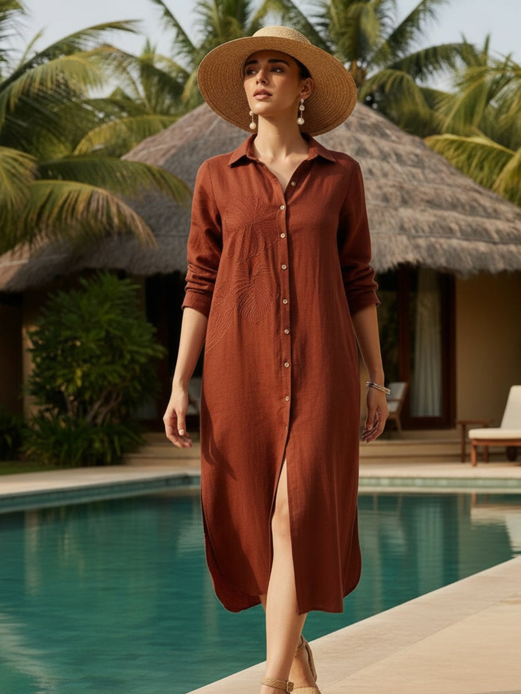 Woman in a rust-colored dress and hat standing by a pool with palm trees in the background