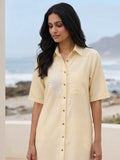 Close up shot of a Woman in a cotton linen blend yellow shirt dress with embroidery holding a straw bag by a beach.