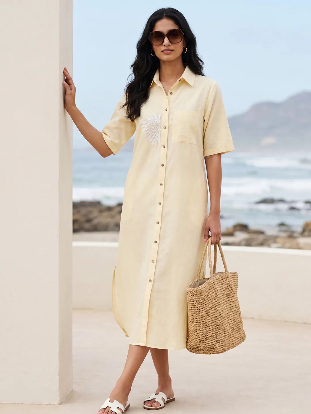 Woman in a cotton linen blend yellow shirt dress with embroidery holding a straw bag by a beach.