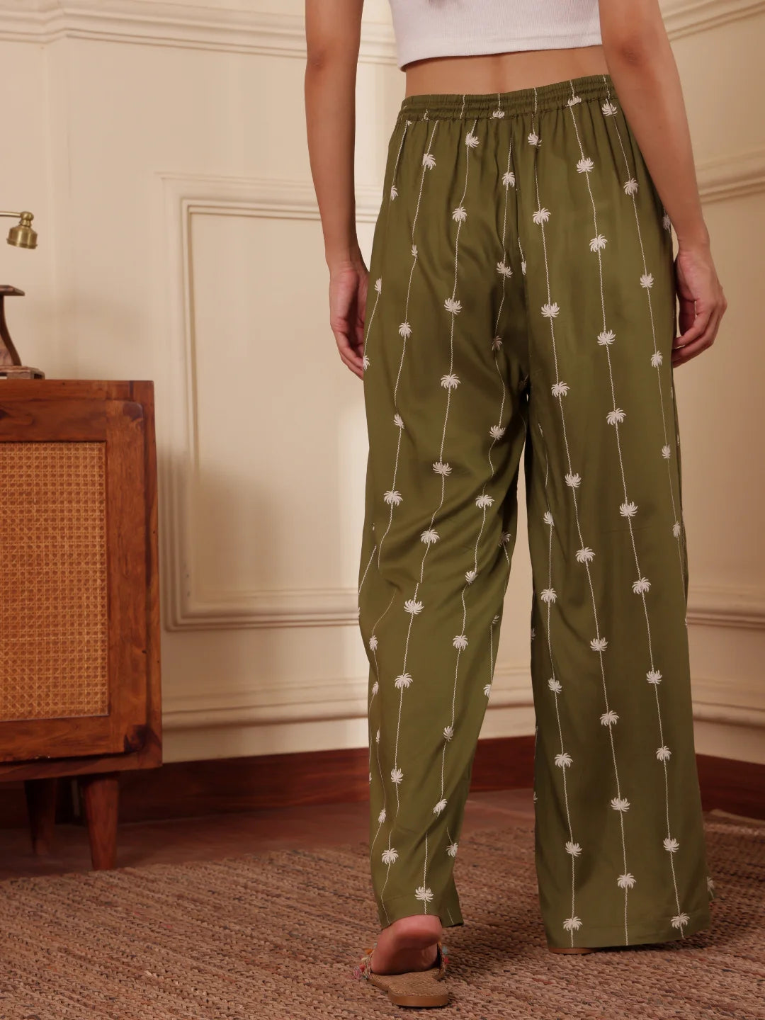 Women back shot in an Olive green Palm tree print pants with drawcord and tassels in a room with wooden furniture.