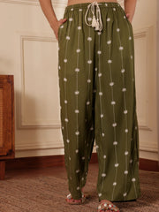 Women walking shot in an Olive green Palm tree print pants with drawcord and tassels in a room with wooden furniture.
