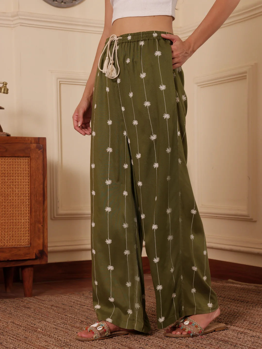 Women walking shot in an Olive green Palm tree print pants with one hand in pocket in a room with wooden furniture.