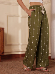 Women right profile shot in an Olive green Palm tree print pants with drawcord and tassels in a room with wooden furniture.