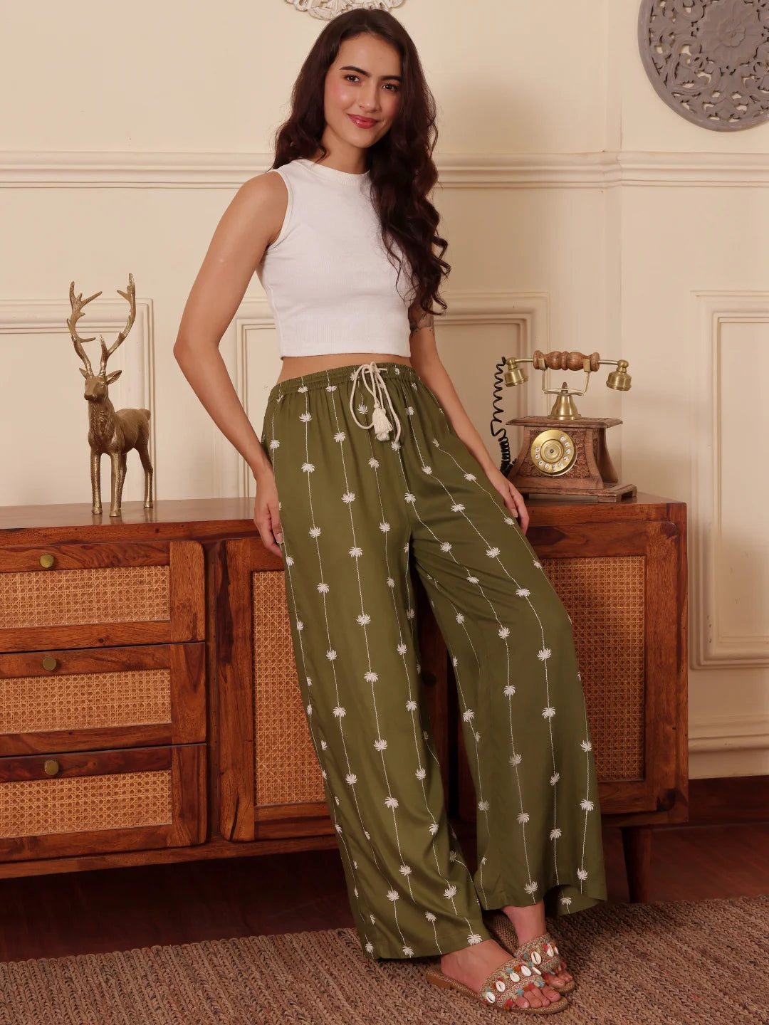 Women leaning against a wooden chest in an Olive green Palm tree print pants with drawcord and tassels in a room 