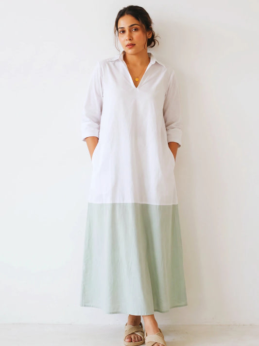 A woman modeling a long white tunic dress with a light sage green skirt section, standing against a plain white wall.