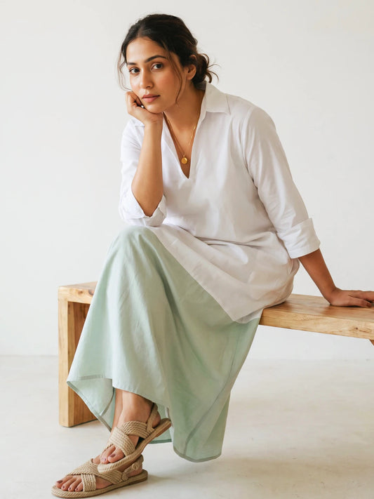 A woman sits on a wooden bench wearing a loose white long-sleeve dress, paired with beige woven sandals, against a plain white wall.