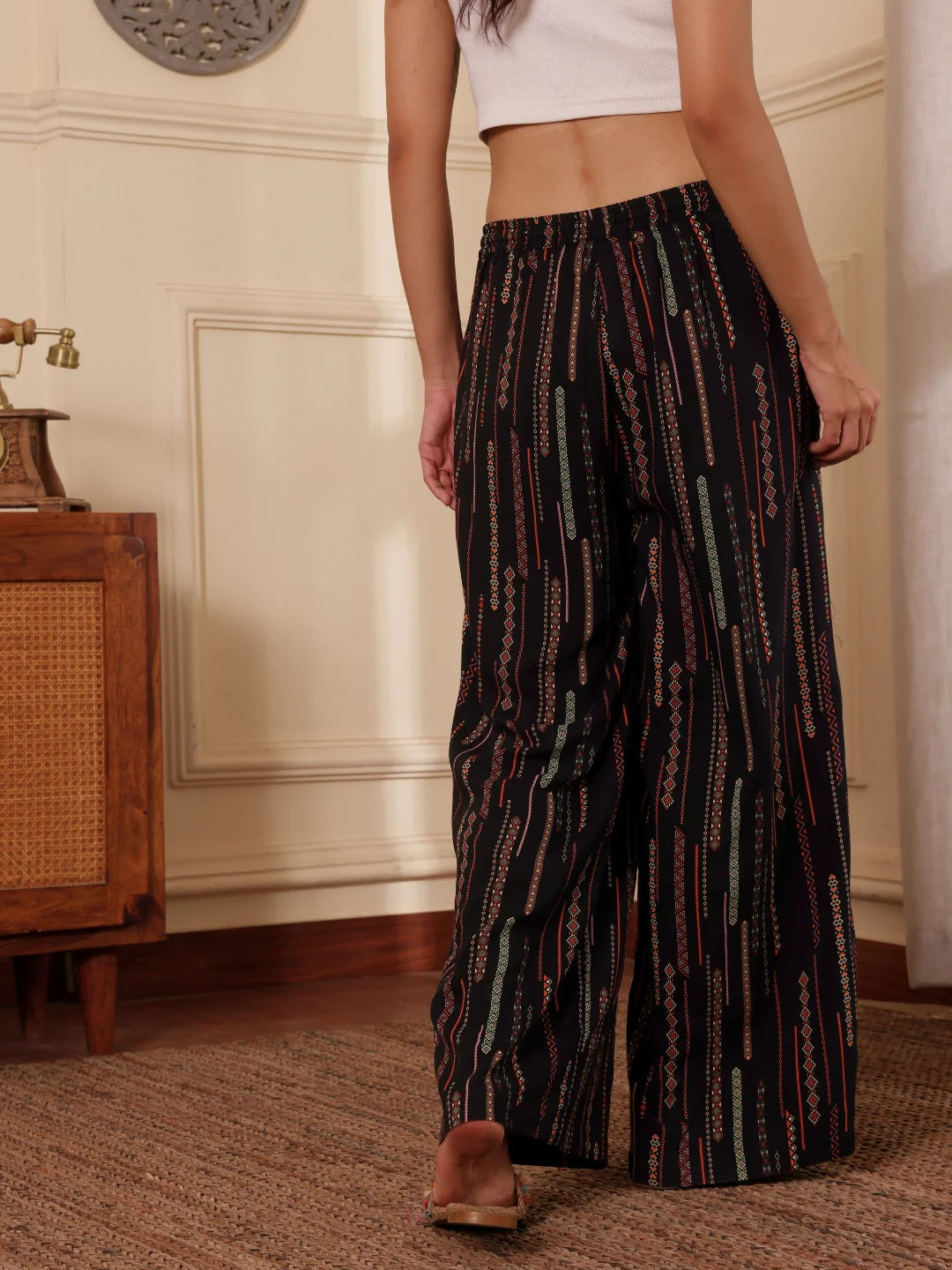 women back shot wearing a black boho print wide leg resort pants with white tank top in a room with textured wall