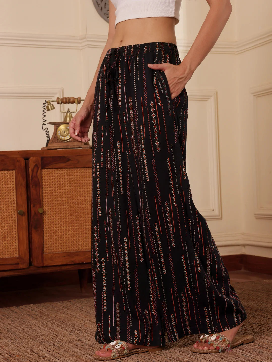 women walking side ways in a black boho print wide leg resort pants with white tank top in a room with textured wall