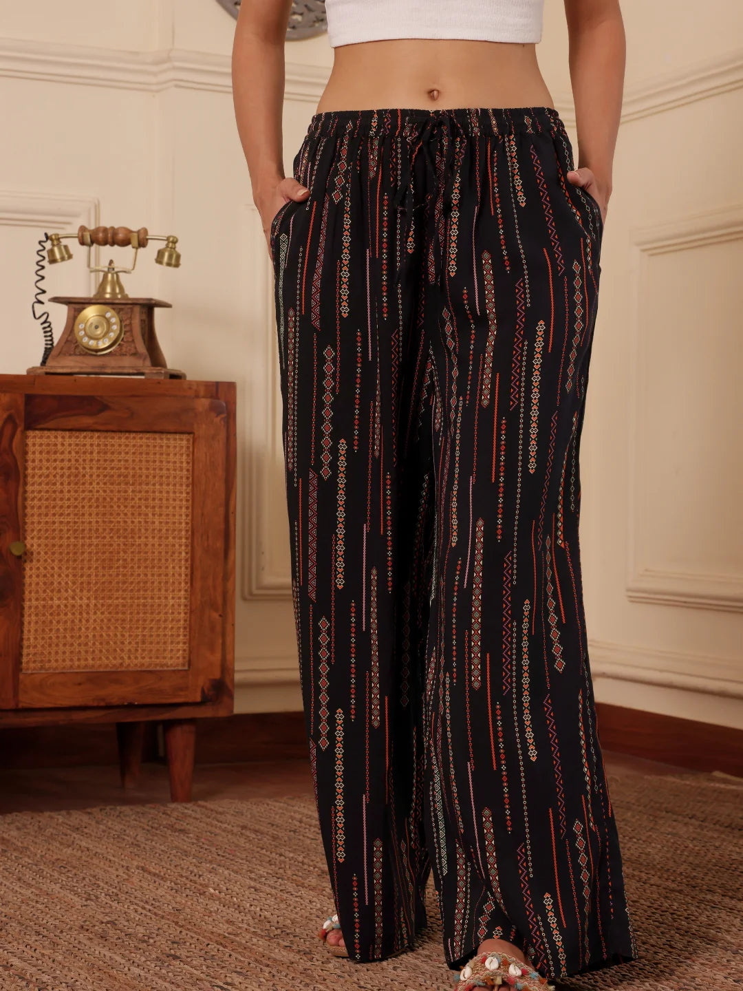 A women standing wearing a black boho print wide leg resort pants with white tank top and both hands inside pocket in a room with textured wall