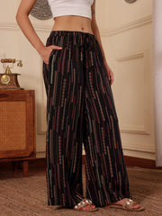women wearing a black boho print wide leg resort pants with white tank top in a room with textured wall