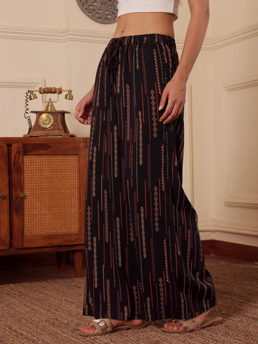 women standing side ways in a black boho print wide leg resort pants with white tank top in a room with textured wall