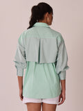A woman shown from the back wearing a mint green and white vertically striped shirt with solid pale green shoulder panels and a large back pleat detail, paired with white shorts against a plain light beige wall.