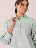 A woman wearing a long-sleeve button-up shirt with a light sage green body and green-and-white striped yoke on the collar and shoulders, posed against a soft pink studio background.