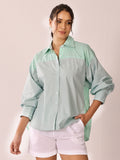 A woman modeling a long-sleeve button-up shirt with a sage green body and mint green and white vertical stripes on the yoke and side panels, paired with white shorts against a light pink background.