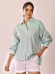 A woman modeling a long-sleeve button-up shirt with a sage green body and mint green and white vertical stripes on the yoke and side panels, paired with white shorts against a light pink background.