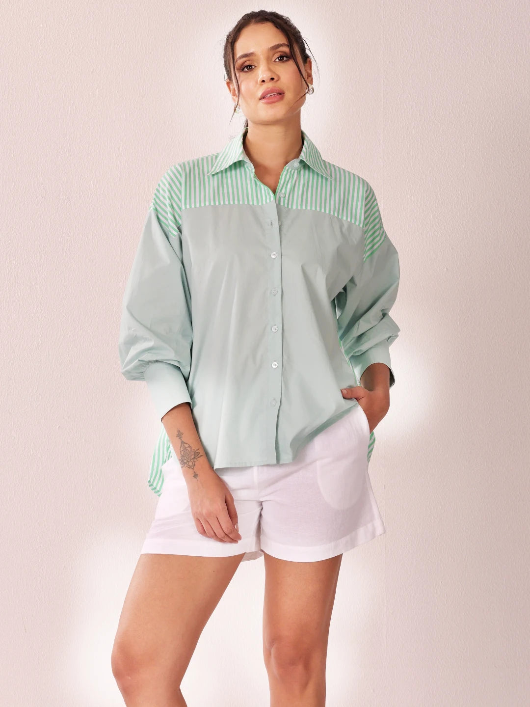 A woman modeling a mint green and white striped button-down shirt with a solid pale green body, paired with white shorts, against a soft pink studio background.