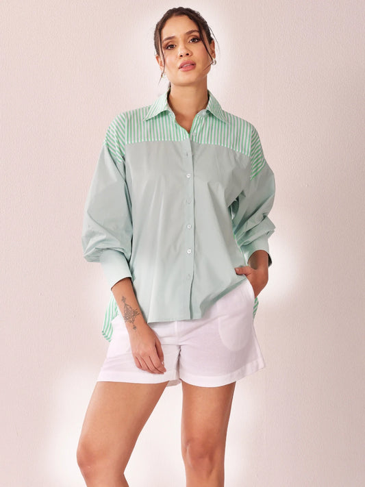 A woman modeling a mint green and white striped button-down shirt with a solid pale green body, paired with white shorts, against a soft pink studio background.