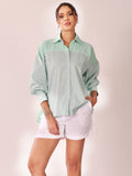 A woman modeling a mint green and white striped button-down shirt with a solid pale green body, paired with white shorts, against a soft pink studio background.