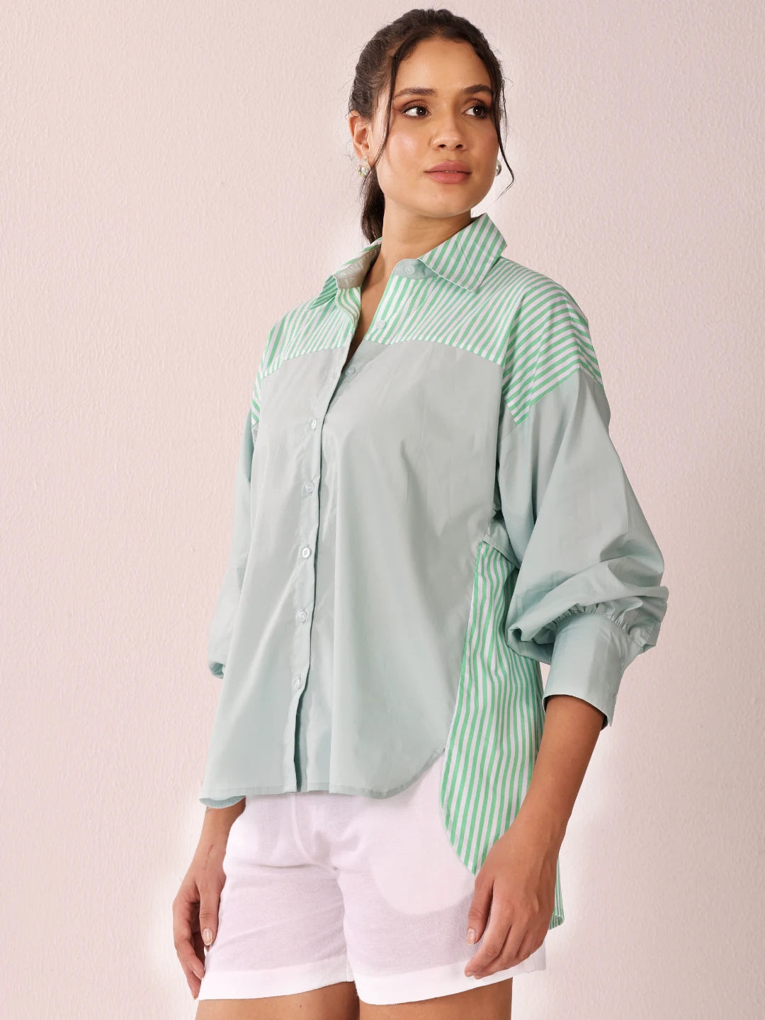 A woman modeling a light sage green button-up shirt with green and white striped panels on the chest, shoulders, and side, paired with white shorts against a pale pink background.