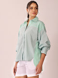 A woman modeling a light sage green button-up shirt with green and white striped panels on the chest, shoulders, and side, paired with white shorts against a pale pink background.