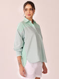 A woman modeling a light sage green oversized button-up shirt with mint green and white striped panels on the shoulders and sides, paired with white shorts against a solid pale pink background.