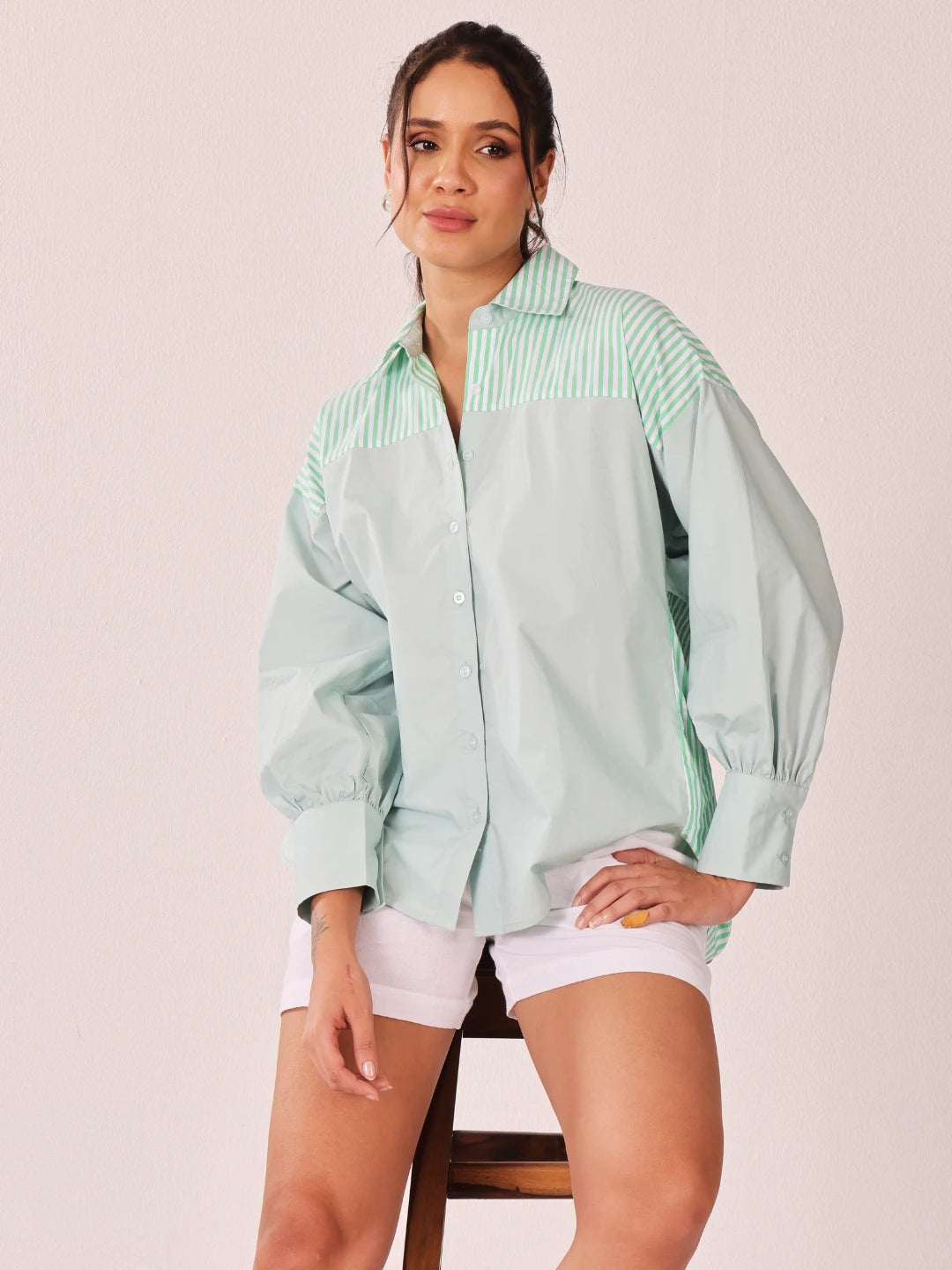A woman models a light mint green color-blocked button-up shirt with a vertically striped yoke, paired with white shorts, seated on a wooden stool against a solid pale pink background.