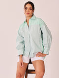 A woman models a light mint green color-blocked button-up shirt with a vertically striped yoke, paired with white shorts, seated on a wooden stool against a solid pale pink background.