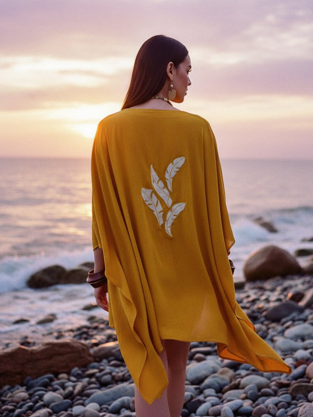 Woman wearing a yellow cover-up with a leaf design on a rocky beach at sunset.