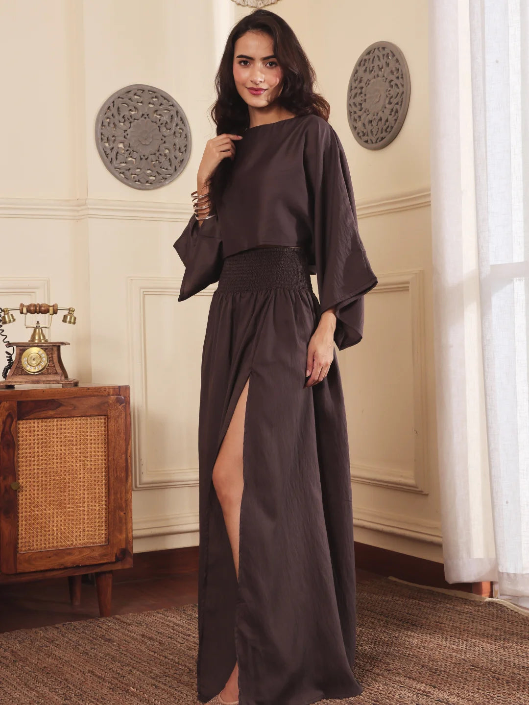 Left pose of a Woman wearing a brown co-ord set with crop top and Skirt with Slit in a room with decorative elements.