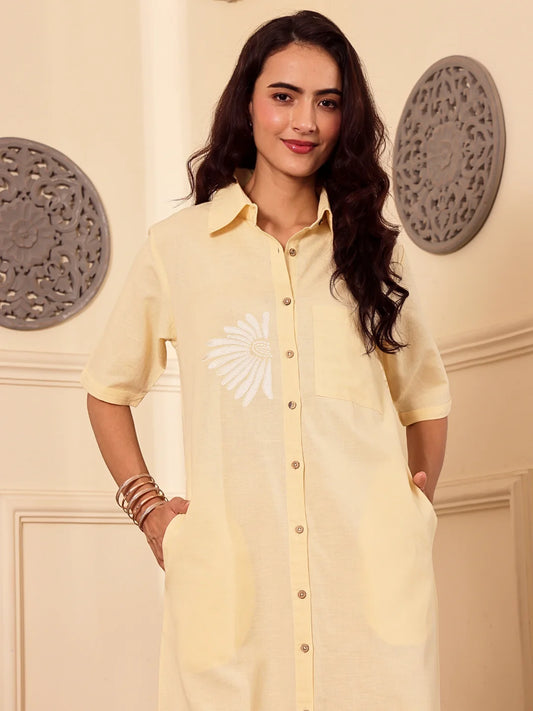 Woman wearing a light yellow cotton linen shirt dress with a floral design in a room with decorative elements.
