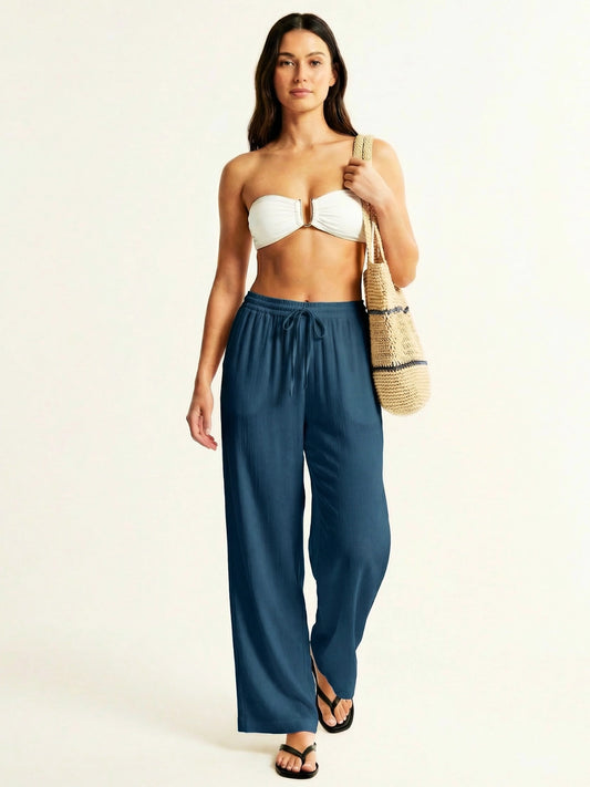 Woman wearing a white strapless top and teal high-waisted resort pants, holding a straw bag on a beige background.