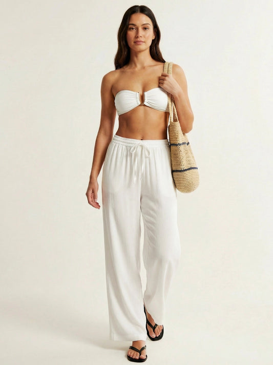Woman wearing a white strapless top and wide-leg pants, holding a straw bag on a plain background