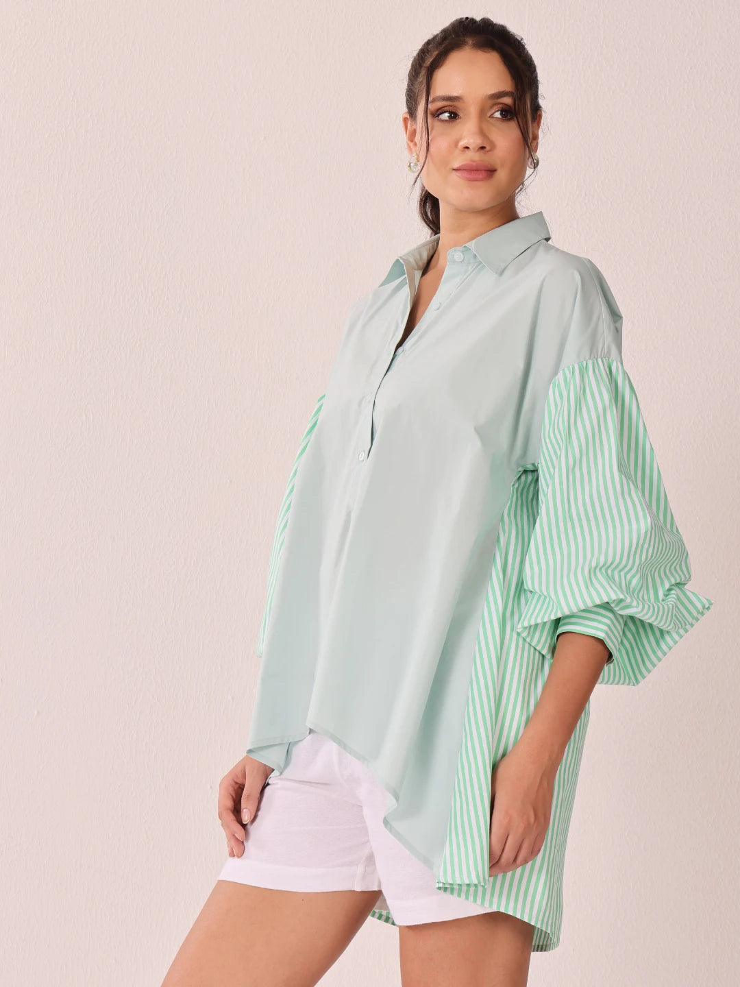 A woman modeling an oversized button-up shirt with a pale mint green body and green-and-white striped bell sleeves, paired with white shorts, against a plain light beige studio background.