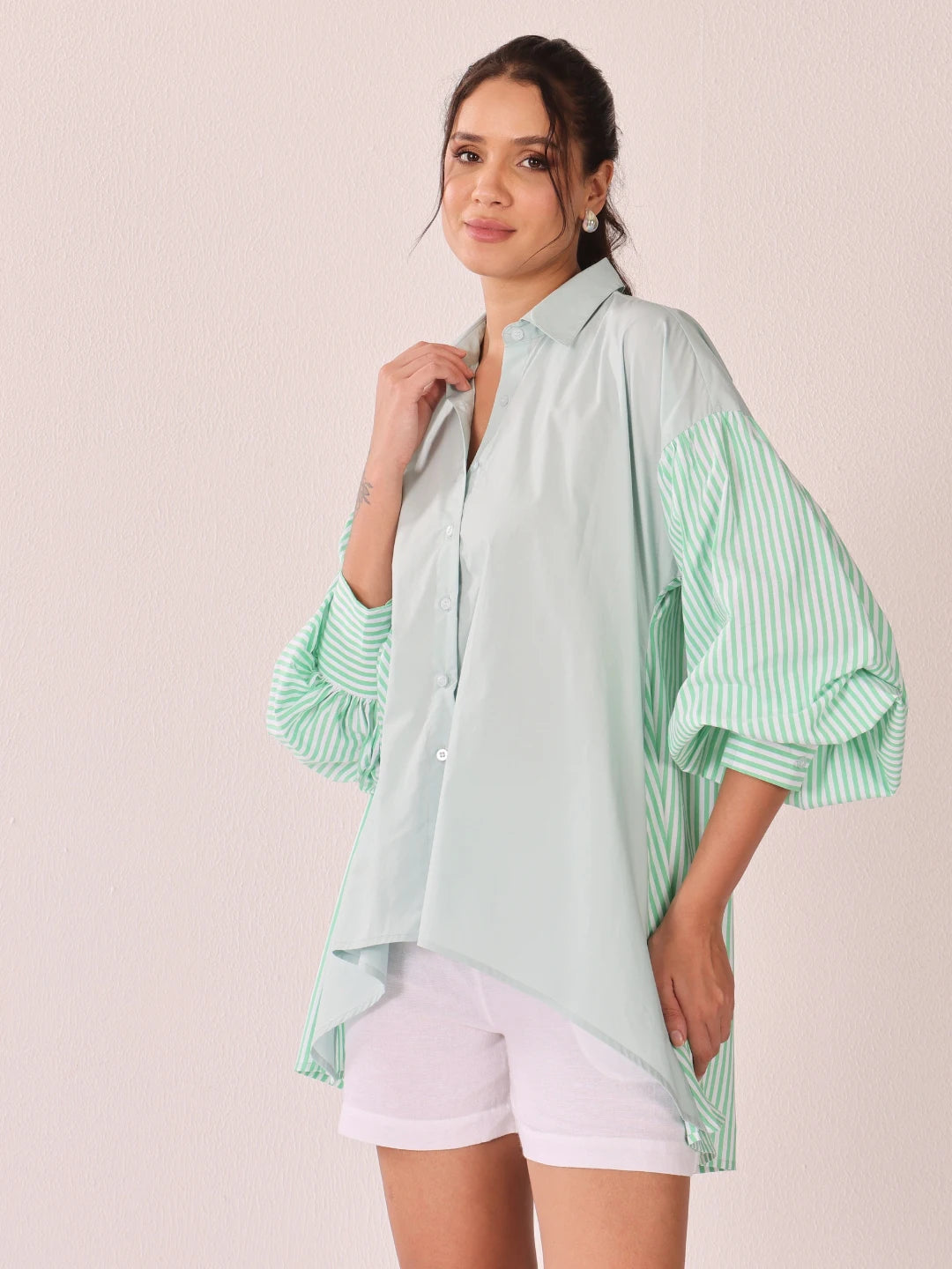 A woman modeling an oversized button-up shirt with a pale mint green body and mint green and white vertical striped sleeves, paired with white shorts against a solid pale pink background.