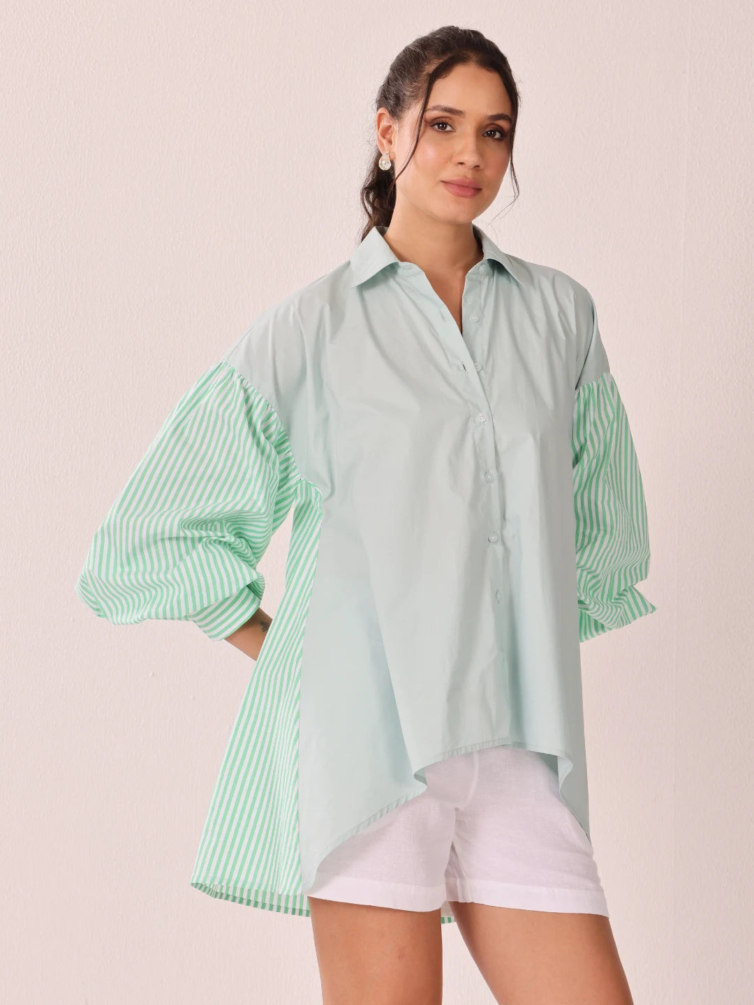 A woman modeling an oversized mint green button-up shirt with contrasting light green and white striped balloon sleeves, paired with white shorts, against a solid pale pink background.