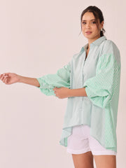 A woman modeling an oversized button-up shirt with mint green and white vertical stripes on the sleeves and a solid pale mint green body, paired with white shorts against a solid light pink background.