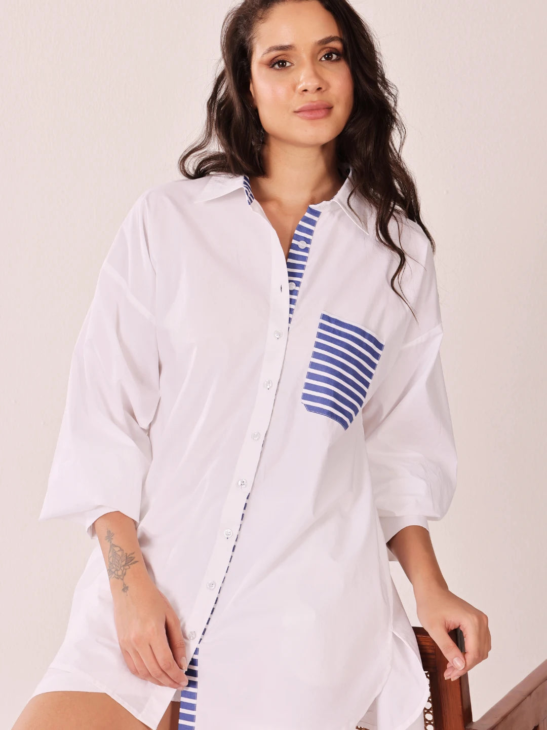 A woman wearing a white oversized button-up shirt dress with blue and white striped accents on the collar, placket, and chest pocket, posed against a light beige background.