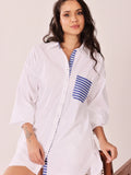 A woman wearing a white oversized button-up shirt dress with blue and white striped accents on the collar, placket, and chest pocket, posed against a light beige background.