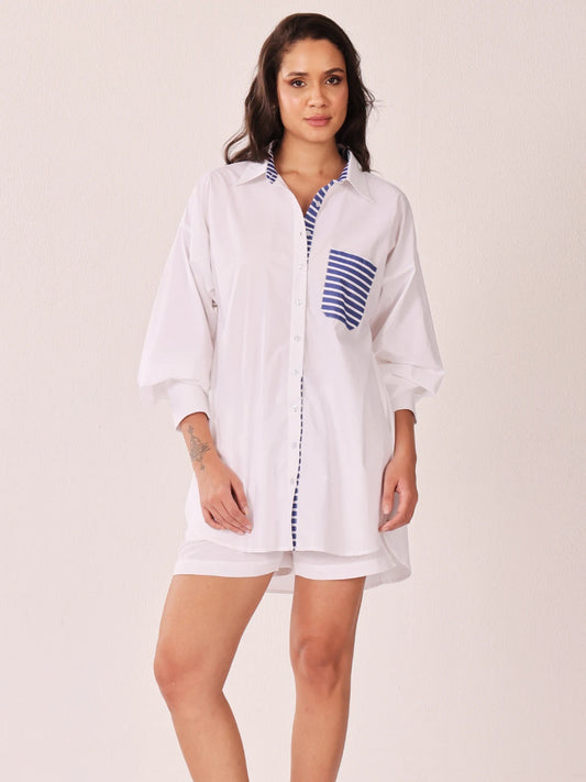 A woman modeling a white button-up shirt dress with blue and white striped accents on the chest pocket and front placket, set against a plain light beige studio background.