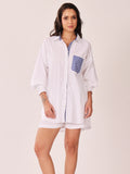 A woman modeling a white button-up shirt dress with blue and white striped accents on the chest pocket and front placket, set against a plain light beige studio background.