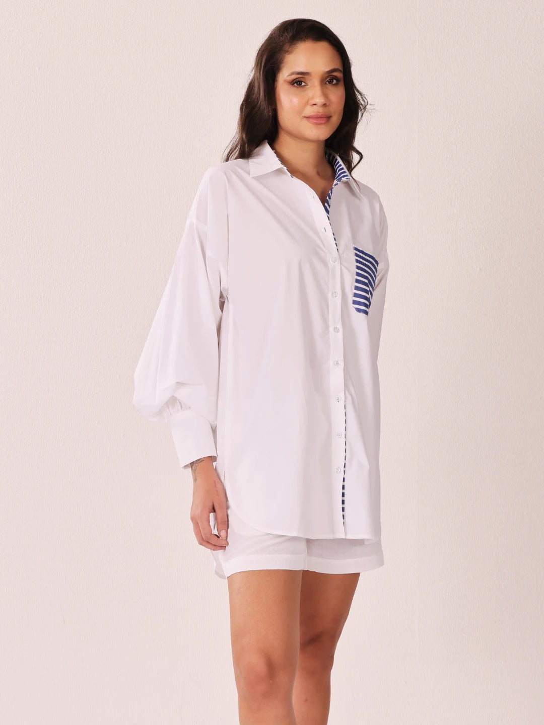 A woman modeling a white oversized button-up shirt with blue and white striped details on the chest pocket and inner collar, paired with matching white shorts, against a plain light beige studio background.