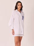 A woman modeling a white oversized button-up shirt with blue and white striped details on the chest pocket and inner collar, paired with matching white shorts, against a plain light beige studio background.