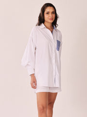 A woman modeling a white oversized button-up shirt with blue and white striped details on the chest pocket and inner collar, paired with matching white shorts, against a plain light beige studio background.