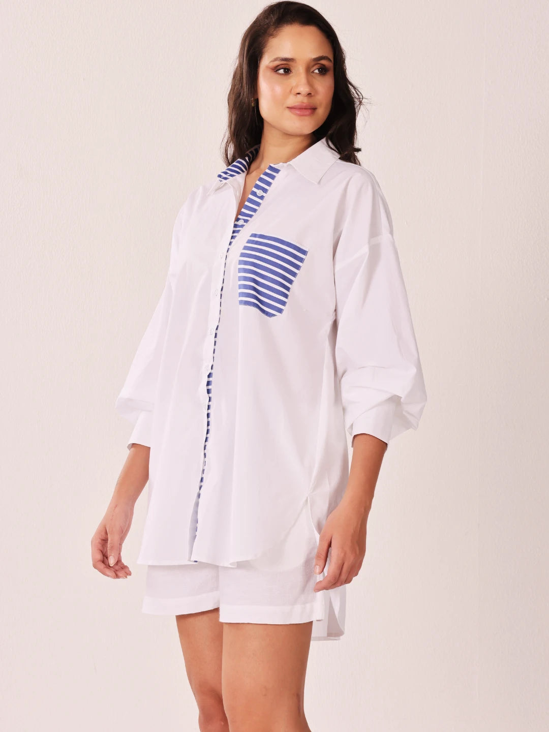 A woman modeling a white oversized button-up shirt dress with blue and white striped accents on the collar, placket, and chest pocket, paired with matching white shorts against a plain light beige studio background.