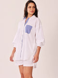 A woman modeling a white oversized button-up shirt dress with blue and white striped accents on the collar, placket, and chest pocket, paired with matching white shorts against a plain light beige studio background.