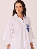 A woman wearing a white oversized button-up shirt with blue striped accents on the pocket and inner collar, photographed against a plain light background.